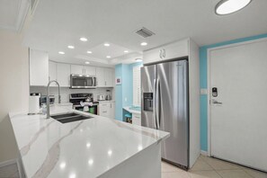 A modern kitchen with white cabinetry, a stainless steel refrigerator, a microwave, and a sink with a marble countertop. - Crystal Sands Tower Unit 704 - Beachfront (Sarasota)
