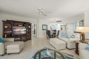 A living room with a TV, a ceiling fan, a dining area with chairs, and a kitchen in the background. - Crystal Sands Tower Unit 704 - Beachfront (Sarasota)