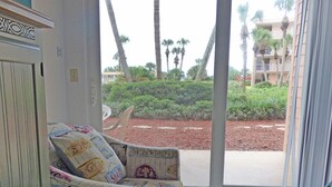 A room with a patterned armchair, a door with decorative molding, and a view of a landscaped area with palm trees and a building in the background. - SEASIDE ESCAPE ON ST. AUGUSTINE BEACH - WHERE OLD IS NEW AGAIN!!! (St Augustine Beach)