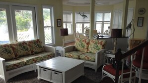 A living room with a white sofa set, a coffee table, and a red chair. There are two windows with blinds, a ceiling fan, and a lamp on a side table.