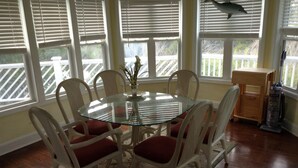 A dining area with a glass table surrounded by wicker chairs with red cushions, a floral rug, and a view of a wooden deck through large windows.