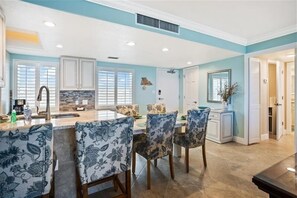 A modern kitchen with a central island, white cabinetry, and a dining area with floral-patterned chairs. - Crystal Sands Tower Unit 710 - Beachfront (Sarasota)