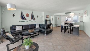A modern living room with a sectional sofa, a dining area with a small kitchen, and a large window. - Crystal Sands Tower Unit 805 - Beachfront (Sarasota)