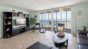 A living room with a TV, bookshelf, and seating area with a view of the ocean. - Crystal Sands Tower Unit 805 - Beachfront (Sarasota)