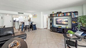 A modern living room with a black leather sofa, a dining area with a white table, and a kitchen in the background. - Crystal Sands Tower Unit 805 - Beachfront (Sarasota)