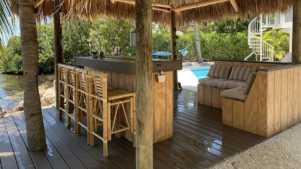 Une terrasse en bois surmontée d’un toit de chaume, un coin bar avec des tabourets, une piscine et un espace détente avec des coussins.