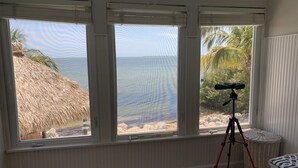 Une pièce dotée de trois grandes fenêtres donnant sur une plage, un télescope monté sur un trépied et un panier en osier.