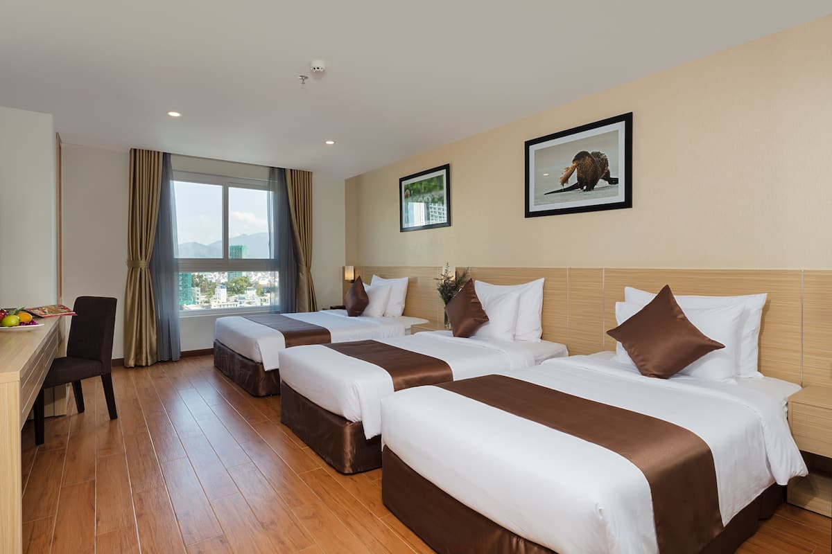 A hotel room with four beds, a desk, and a chair. There is a window with a view of buildings and mountains.