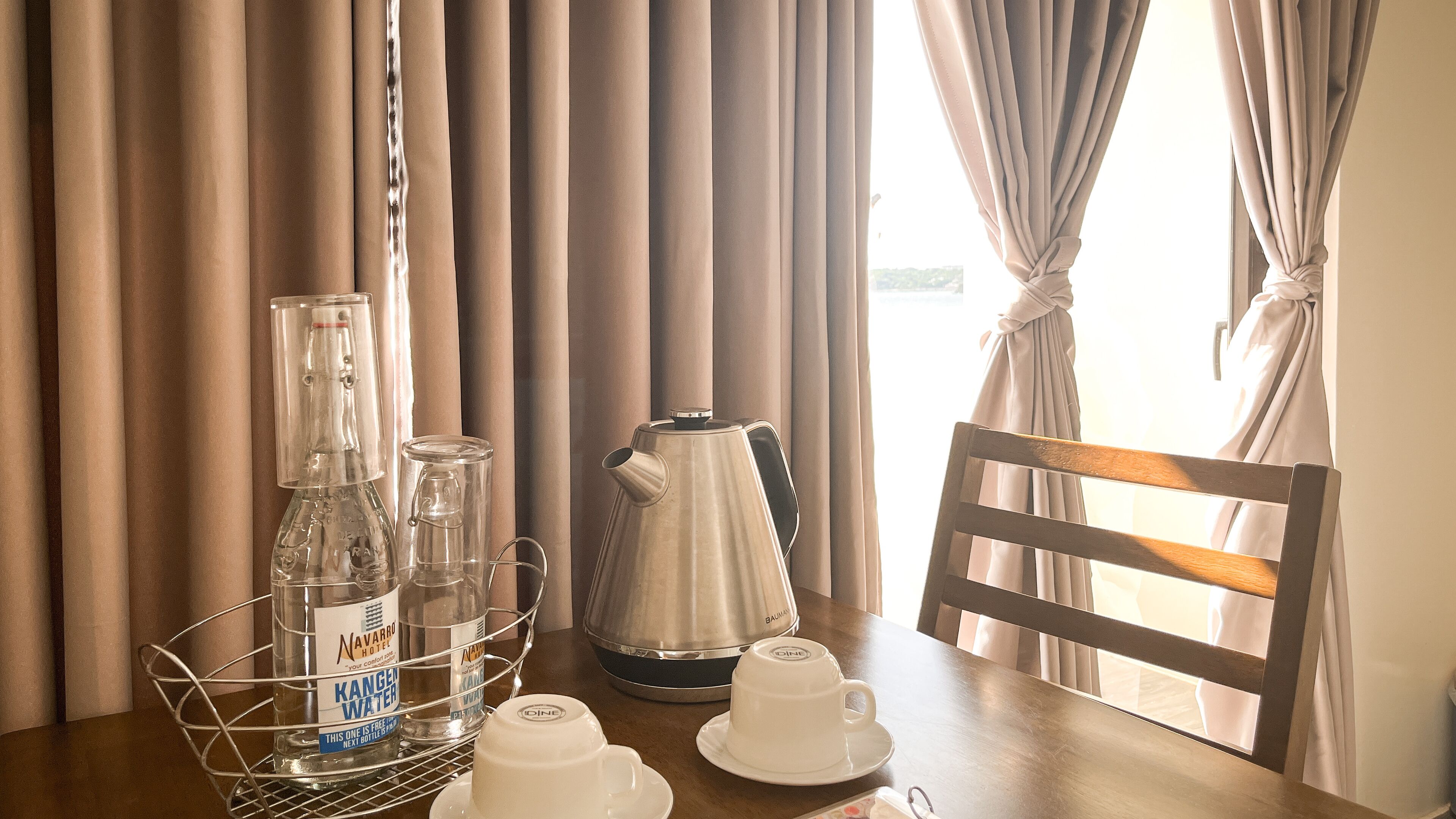 A hotel room with a wooden table set for breakfast, including a bottle of Kangen water, a coffee pot, and white cups.