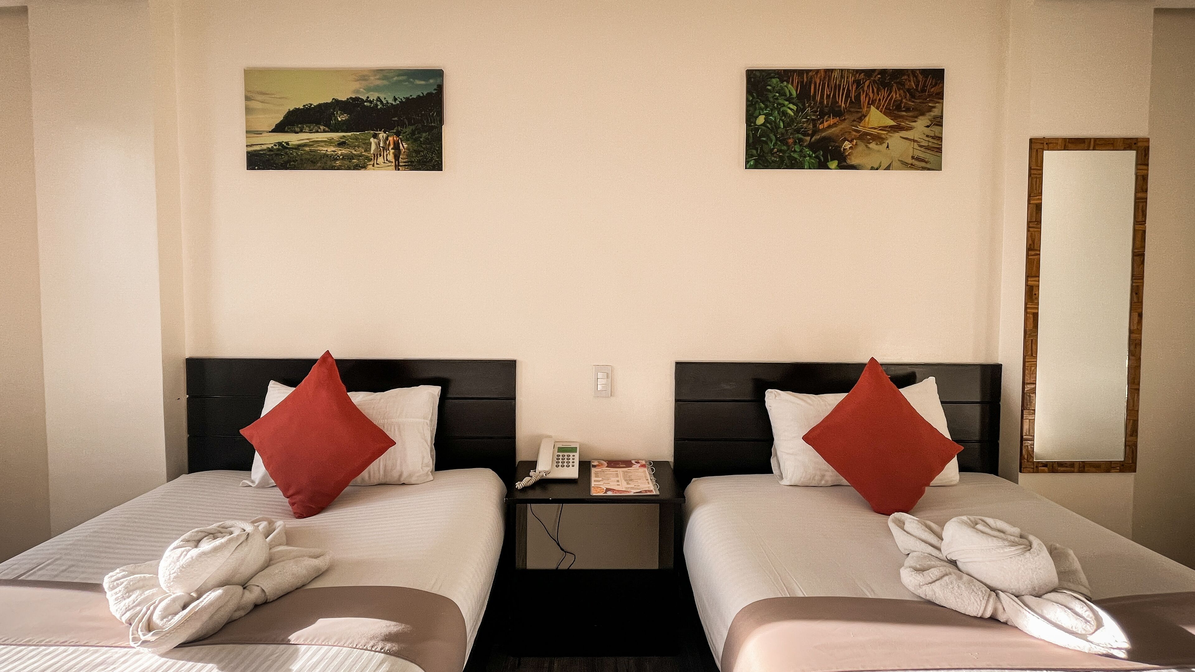 Ein Hotelzimmer mit zwei Betten, jedes mit einem roten Kopfkissen und einem Handtuch. Dazwischen befinden sich zwei Nachttische, auf denen ein Telefon und eine Zeitschrift liegen. An der Wand hängen zwei Bilder und auf der rechten Seite ein Spiegel.