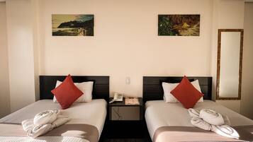 Ein Hotelzimmer mit zwei Betten, jedes mit einem roten Kopfkissen und einem Handtuch. Dazwischen befinden sich zwei Nachttische, auf denen ein Telefon und eine Zeitschrift liegen. An der Wand hängen zwei Bilder und auf der rechten Seite ein Spiegel.