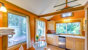 A compact kitchen with wooden cabinets, a stainless steel dishwasher, and a window overlooking a forest. - Pet Friendly - Peaceful Cabin in the Wood - Hot Tub (Bainbridge Island)