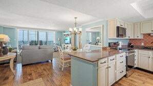 A spacious living room with a grey sofa, a dining area with a round table, and a kitchen with white cabinets and a granite countertop. - Crystal Sands Tower Unit 1204 - Beachfront (SARASOTA)