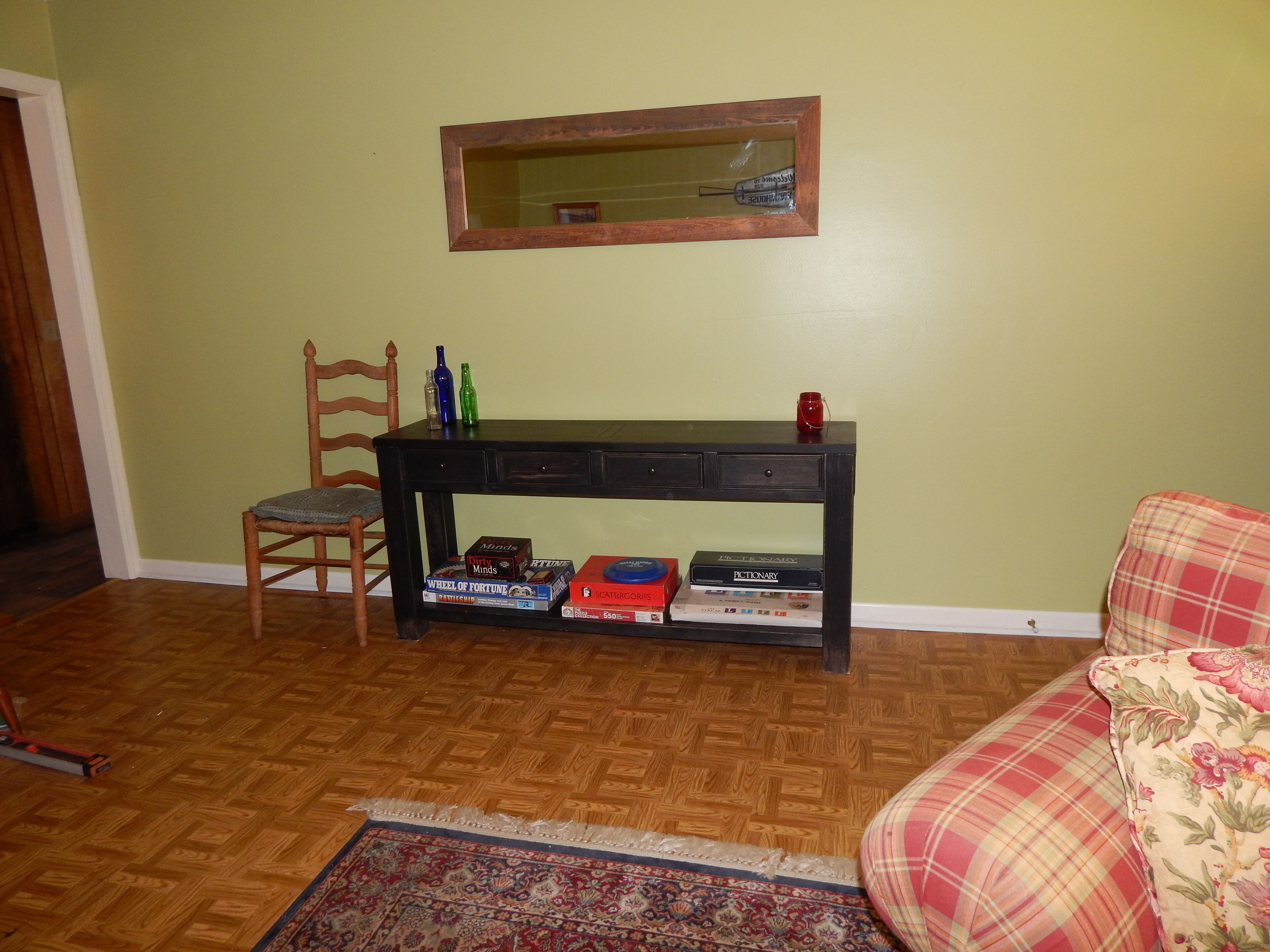 A living room with a wooden floor, a plaid sofa, a wooden chair, a black console table with books, and a mirror on the wall.