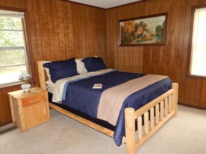 A wooden paneled bedroom with a bed, a nightstand, a lamp, and a painting on the wall. - Hillbilly Hut of South Georgia  (Pitts)
