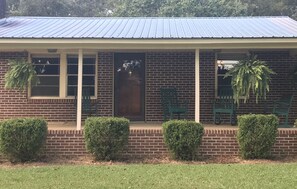 A two-story brick house with a metal roof, a front porch with green chairs, and a large potted plant on the right. - Hillbilly Hut of South Georgia  (Pitts)