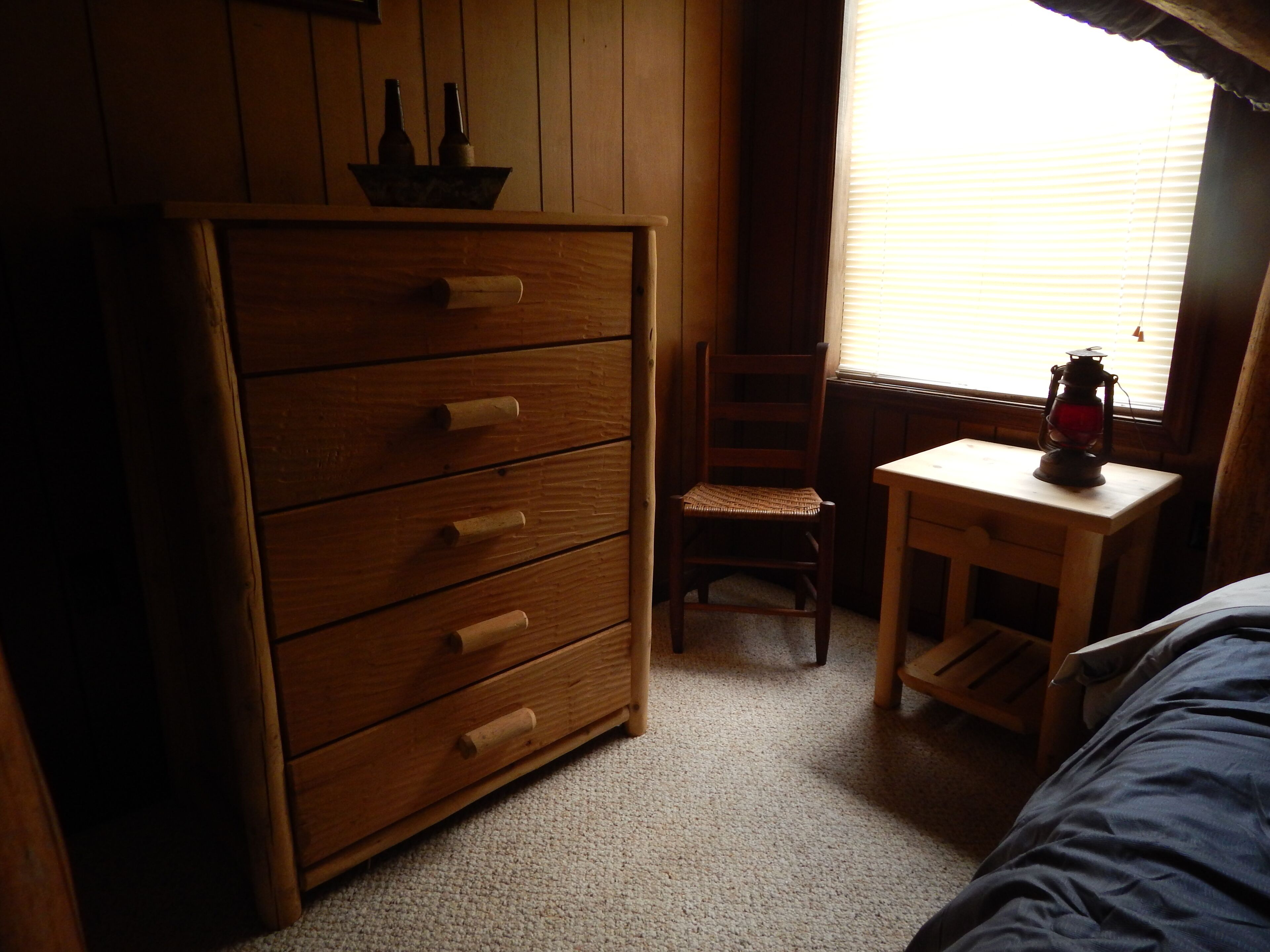 Una habitación con un aparador de madera, una silla, una mesita con un farolillo y una ventana con persianas.