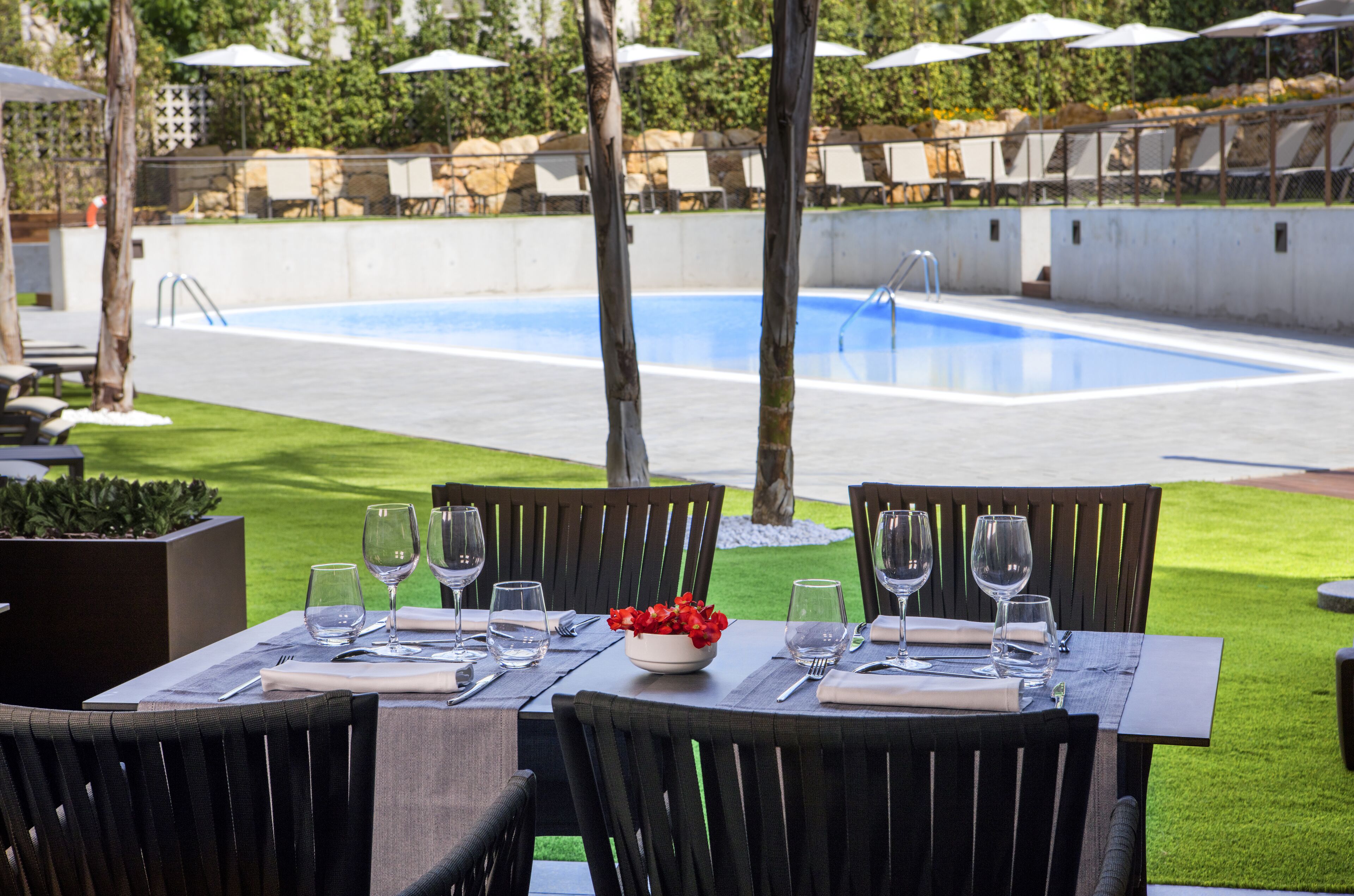 Un área de comedor al aire libre con vista a la piscina, mesas dispuestas con copas de vino y un cuenco con flores rojas.