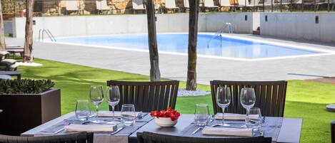 Un área de comedor al aire libre con vista a la piscina, mesas dispuestas con copas de vino y un cuenco con flores rojas.