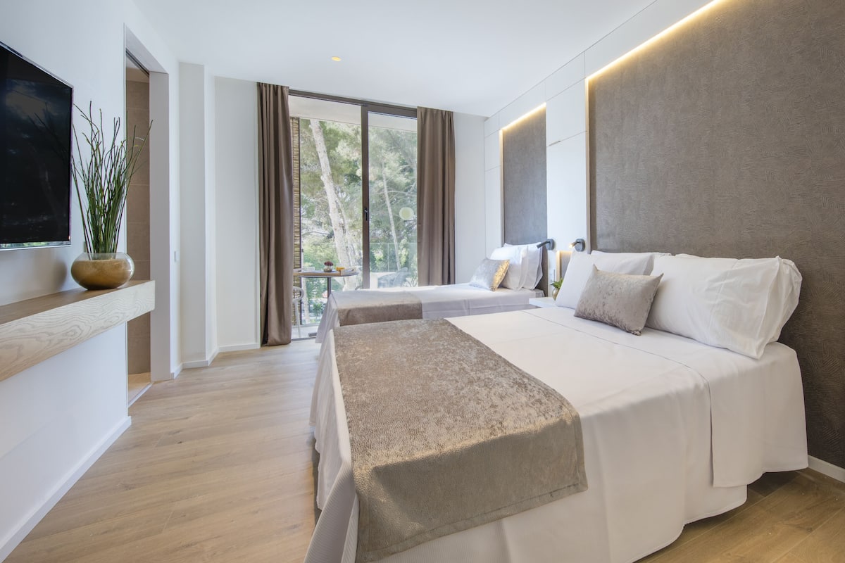 Une chambre d’hôtel moderne avec deux lits, une grande fenêtre avec des rideaux, un téléviseur à écran plat et une petite table surmontée d’un vase.