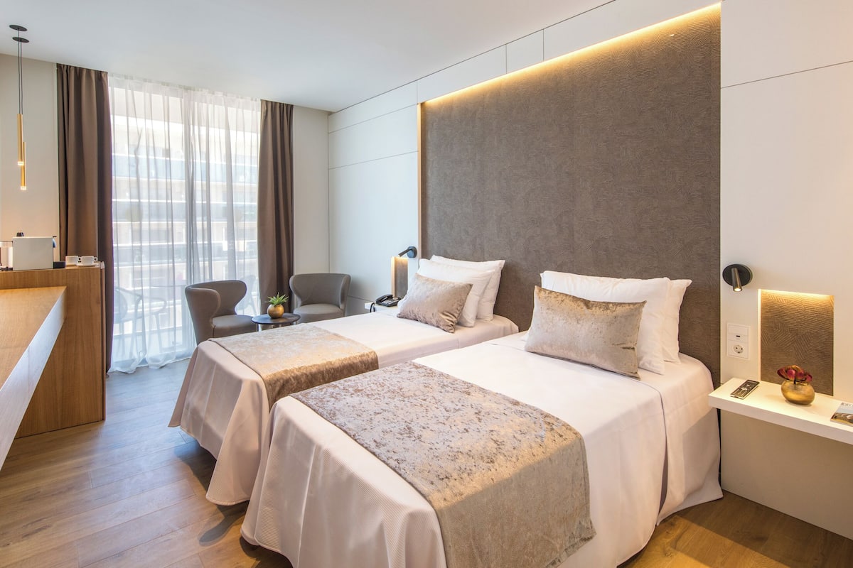 A hotel room with two beds, a desk, a chair, a TV, and a window with curtains.