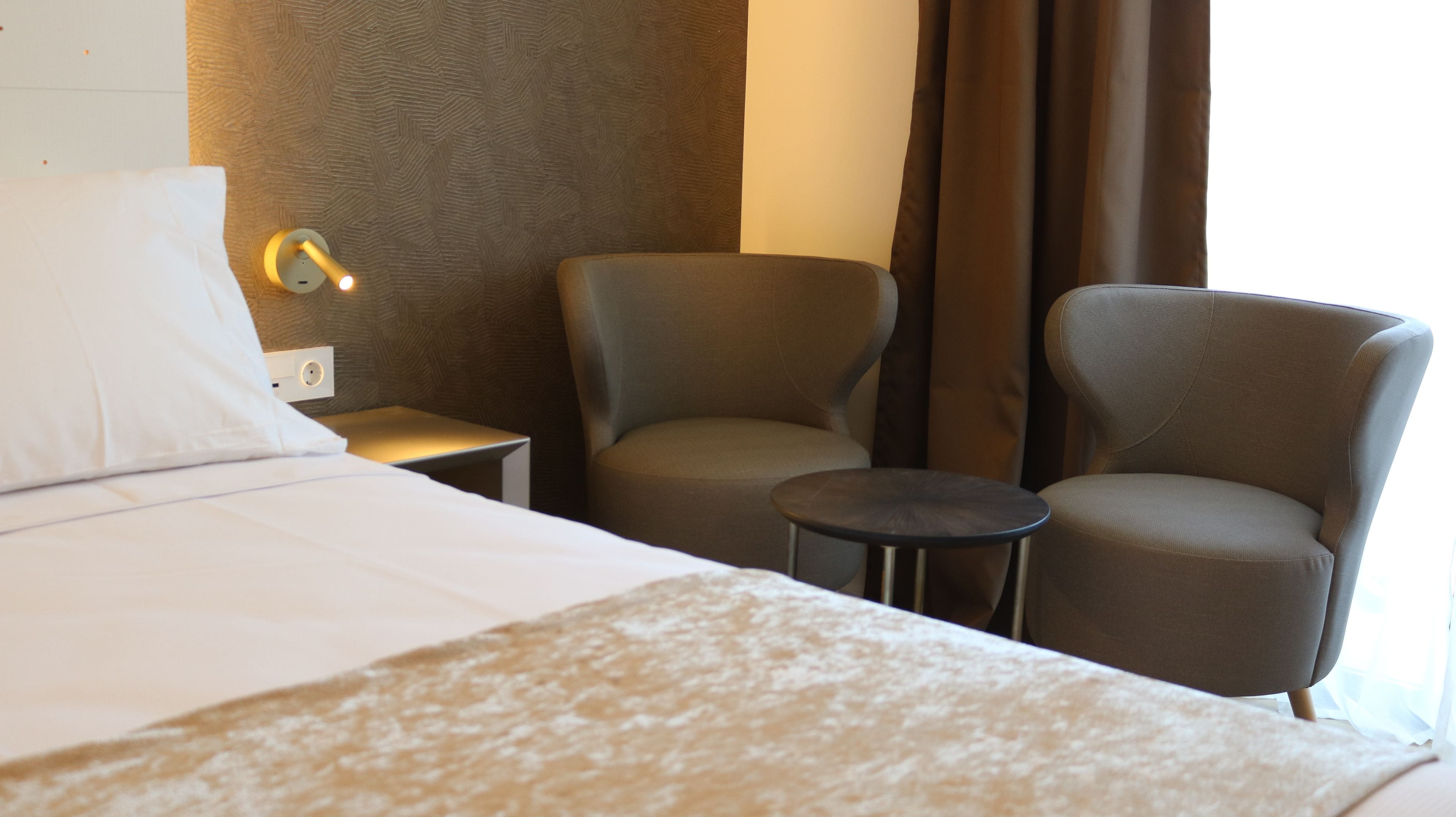 Habitación de hotel con una cama, dos sillones, una mesita y una lámpara en la pared.