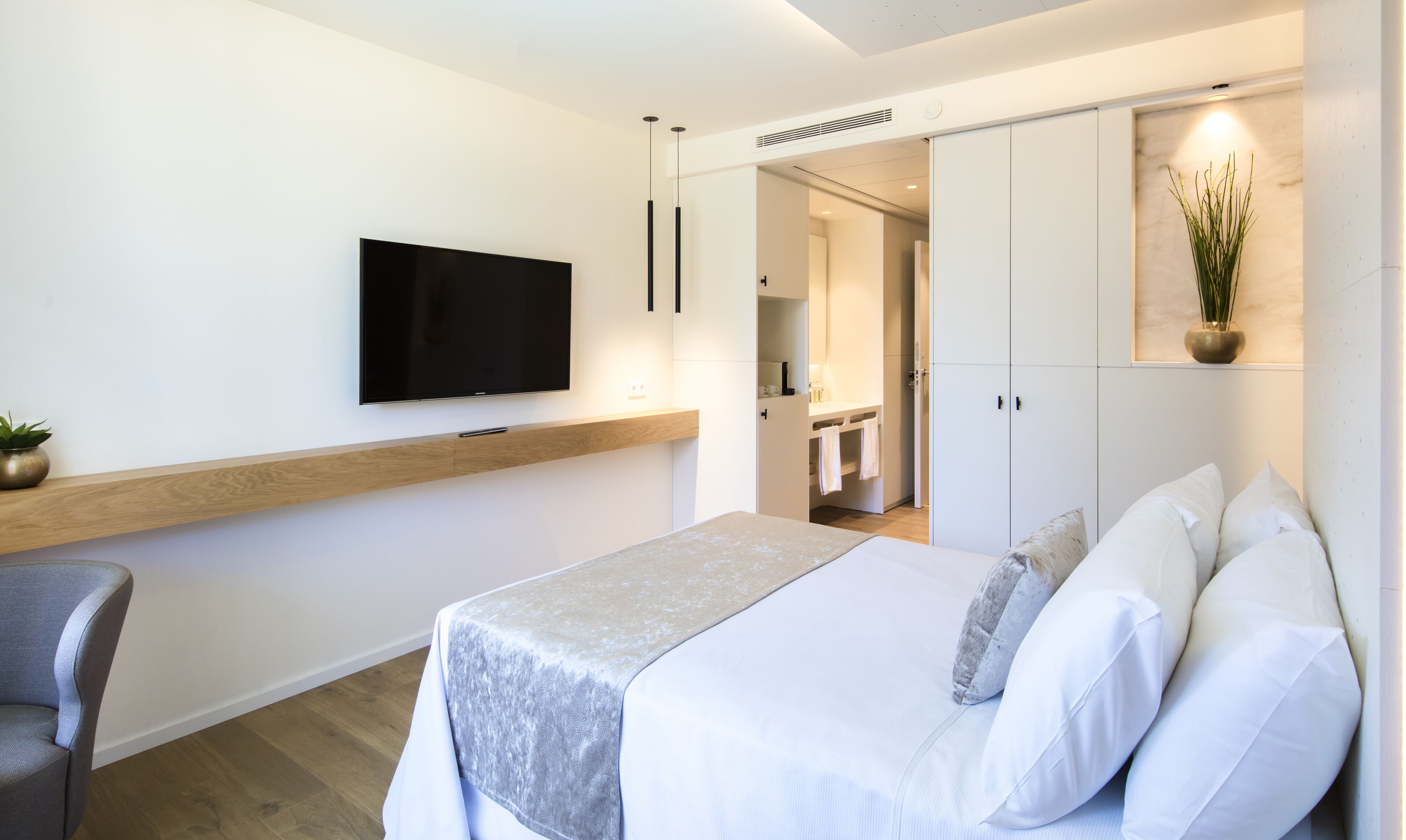Habitación de hotel moderna con una cama, un televisor montado en la pared, un estante de madera y una planta.