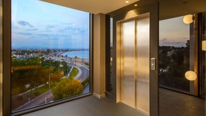 Un ascenseur moderne doté d’une grande fenêtre offrant une vue sur un paysage urbain côtier, comprenant une plage et une route bordée de palmiers.
