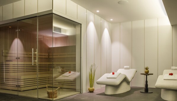 A modern spa room with a glass-enclosed sauna, two massage chairs, and a small table with a plant. - Golden Costa Salou - Adults Only (Vila-Seca)