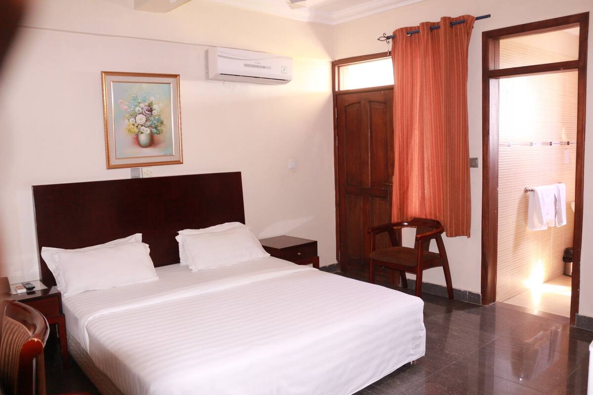 A hotel room with a large bed, a wooden chair, a nightstand, a desk, an air conditioner, a door with an orange curtain, and a framed picture on the wall.