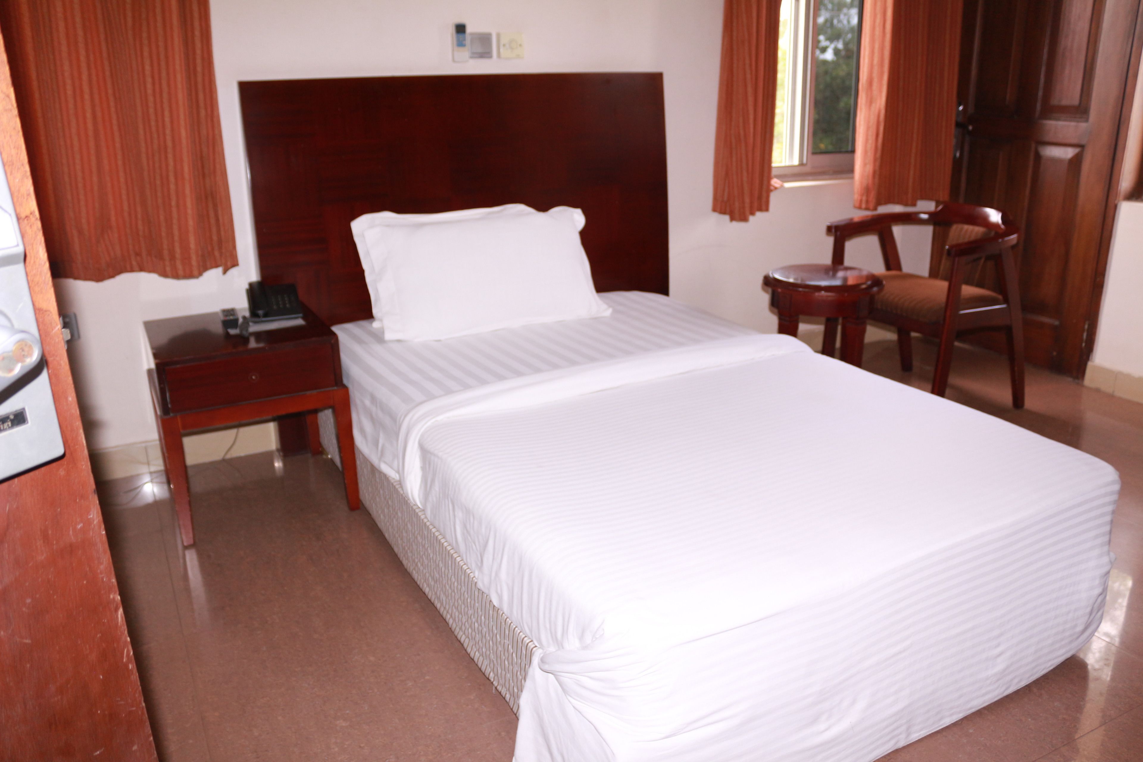 A neatly made bed with a wooden headboard, a wooden nightstand with a phone, and a wooden chair in a hotel room.