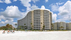 Un hotel grande de varios pisos con fachada curva, situado en una playa con palmeras y tumbonas.