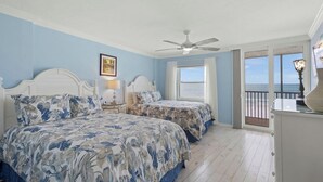 Un dormitorio con dos camas, ventilador de techo, ventana con vista al océano y balcón con barandilla.