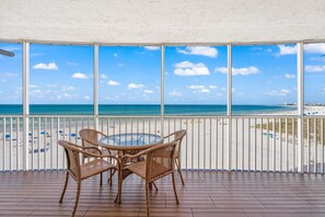 Un balcon avec des fauteuils en osier et une table en verre, donnant sur une plage parsemée de parasols et sous un ciel dégagé.
