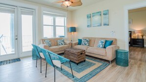 A living room with a beige sofa, a blue and white patterned armchair, a woven coffee table, and a teal side table. There is a ceiling fan, a floor lamp, and artwork on the wall. - Sea Pointe- The Perfect Beachfront Destination - Brand New Elevator (Galveston)