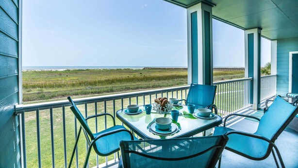 A covered outdoor dining area with a table set for two, overlooking a grassy field and the ocean. - Sea Pointe- The Perfect Beachfront Destination - Brand New Elevator (Galveston)