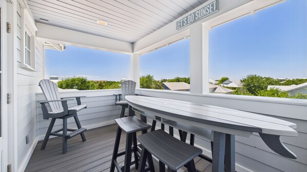 A covered outdoor seating area with a table and chairs, a sign reading "LET'S DO SUNSET", and a view of houses and greenery in the background.
