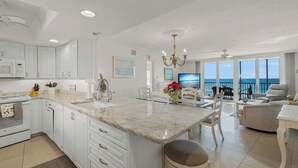 A modern kitchen with white cabinets, a marble island, and a dining area with a view of the ocean. - Crystal Sands Tower Unit 502 - Beachfront (SARASOTA)