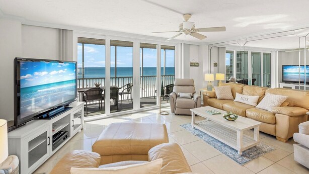 A spacious living room with a large TV, comfortable seating, and a balcony with a view of the ocean. - Crystal Sands Tower Unit 502 - Beachfront (SARASOTA)