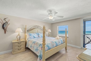 A bedroom with a bed, a ceiling fan, a nightstand with a lamp, a dresser, and a view of the beach through a window.
