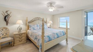A bedroom with a four-poster bed, a ceiling fan, a window with a view of the ocean, and a nightstand with a lamp. - Crystal Sands Tower Unit 305 - Beachfront (Sarasota)