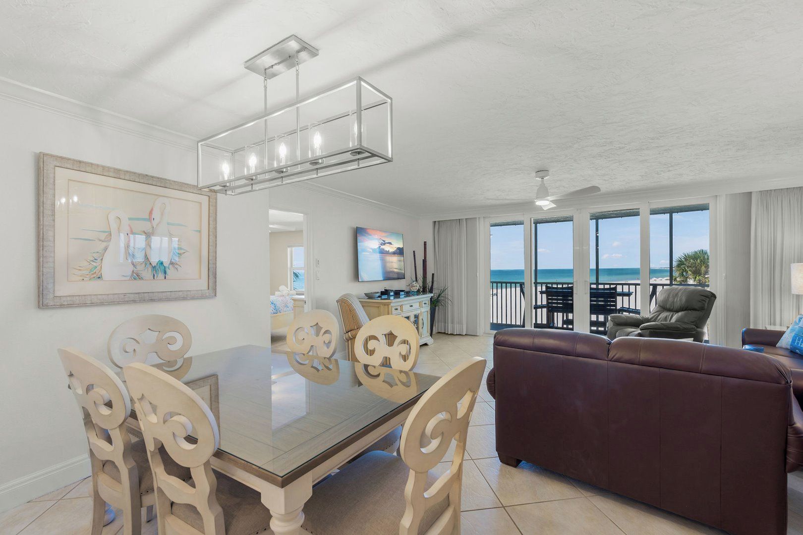 A dining area with a glass table and chairs, a brown leather sofa, a ceiling fan, and a view of the ocean through large windows.