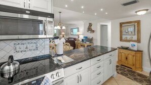 A modern kitchen with a built-in microwave, stove, and oven. There is a dining area with a table and chairs, and a living room with a sofa and a TV. - Crystal Sands Tower Unit 305 - Beachfront (Sarasota)