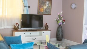 A living room with a flat-screen TV on a wooden stand, a glass coffee table, a blue sofa, a white cabinet, decorative items, and a vase with flowers. - Studio Deluxe Avec Services Hôteliers - Adults Only (Rawai)