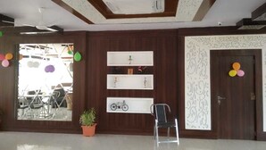 A spacious room with a high ceiling, a ceiling fan, and a built-in air conditioning unit. There are three white shelves against the wall, each with decorative items. A potted plant is on the floor, and a chair is positioned near the shelves. - Rock Star Hotel (Pushkar)