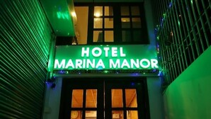 Entrada do Hotel Marina Manor com um letreiro de neon, mostrando uma porta com persiana fechada e uma janela visível.