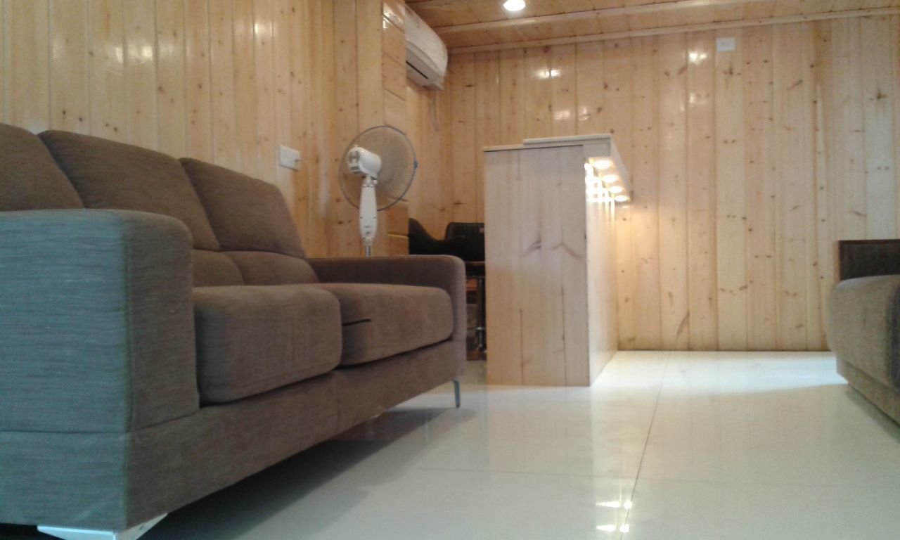 Habitació amb panells de fusta, un sofà, un ventilador de peu i una porta que dona a un altre espai.