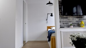 Quarto moderno com cama, televisão montada numa parede de tijolos, escrivaninha com candeeiro e planta em vaso.