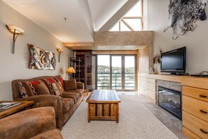 Relax in a spacious living area with a high ceiling, featuring a leather sofa, flat-screen TV, and sliding glass door to a forest-view balcony.