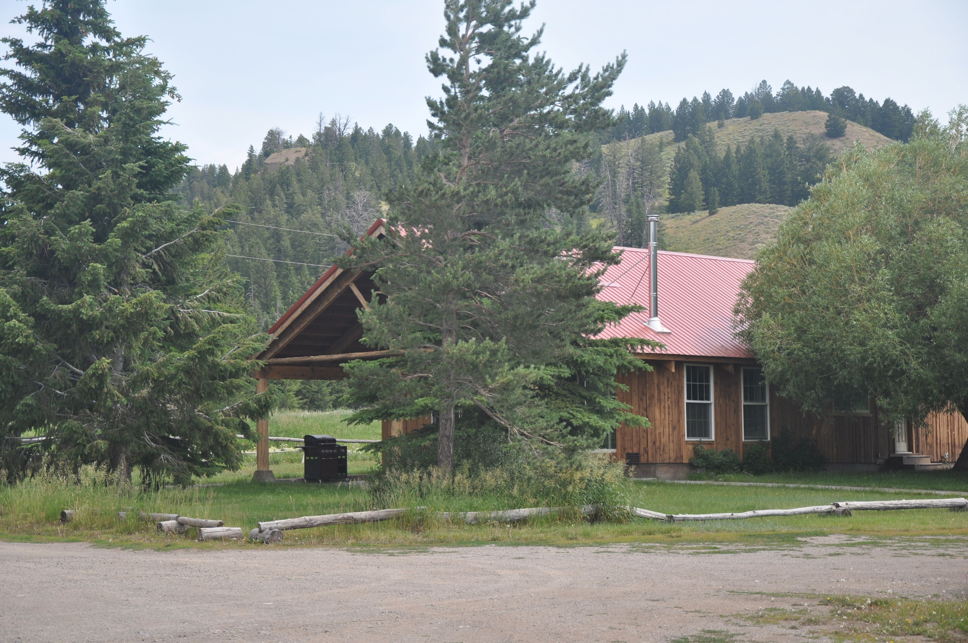 A rustic cabin with a red roof, surrounded by dense forest and hills in the background.